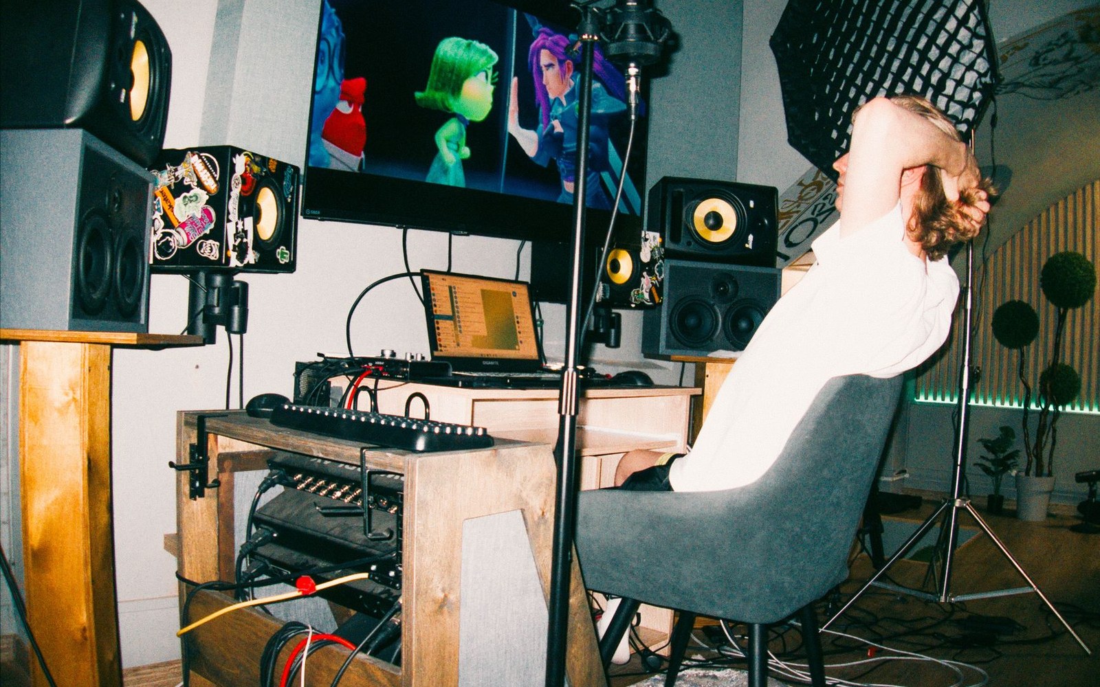 A bedroom studio corner: rack of audio interfaces and studio monitors, adult seated at the position with headphones on, natural window light from the left
