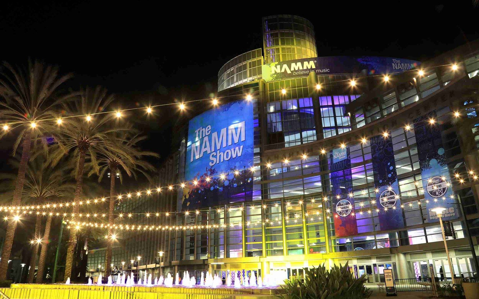 Convention center lit up at night with "The NAMM Show" banner and string lights over palm trees