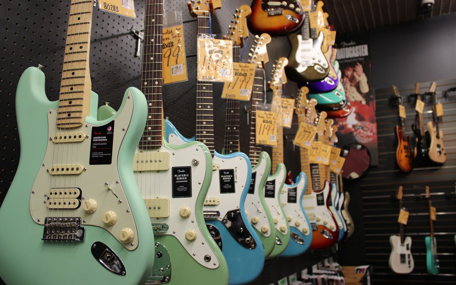 A wall of electric guitars hanging in a shop, photographed straight on, staff member standing to the right at working scale